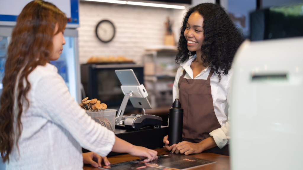 A person making a transaction on a cafe POS system while the staff member helps.