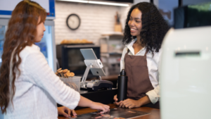 A person making a transaction on a cafe POS system while the staff member helps.